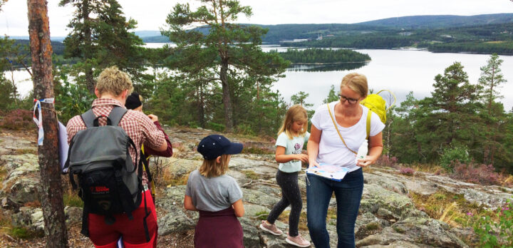 En familj står på ett berg med vacker utsikt över skog och hav, och ska börja gå en stig. Foto.