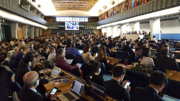 En stor plenum-sal i en internationell förhandlinssituation, full med folk som sitter i svängda bänkrader, med ett podium längst till vänster där en panel sitter. Foto.