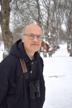 Porträttbild av en man utomhus i lätt snöväder, med en kikare om halsen. Foto.