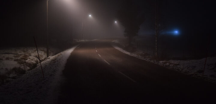 Landsväg en mörk natt, upplyst av gatlyktor. Långt bort kommer en bil. Foto.