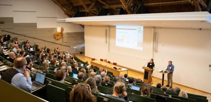 En föreläsningssal med sluttande gradäng, full med folk. Två personer står framme vid podiet. Foto.