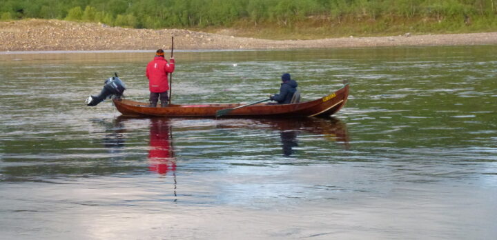 Två personer i en smal båt med en båtmotor. En av personerna håller i en slags pinne. Two persons in a narrow boat with a motor. One of them is holding some sort of stick in the hand. Photo.