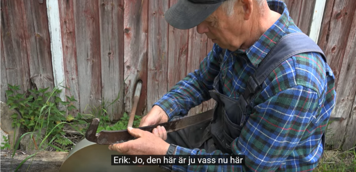 Bild ur en video där man ser en äldre man sitta vid en slipsten och pröva eggen på en lie. Foto.