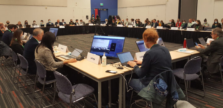 I ett stort rum med kala väggar, ett konferensbord i sluten fyrkant med ett sextiotal människor som sitter vid sina datorer. Foto.