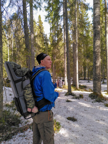 En person står i en vinterskog. På ryggen en bärstol med ett litet barn i. Foto.