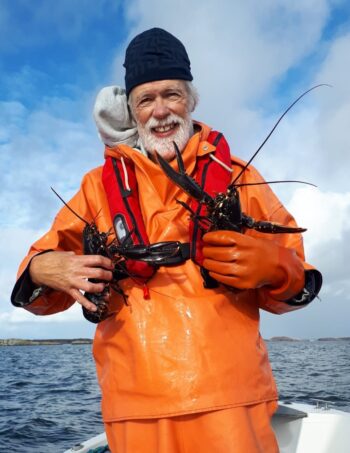 En man i orangea regnkläder i en båt på öppna havet, håller i ett par stora kräftdjur. Foto.